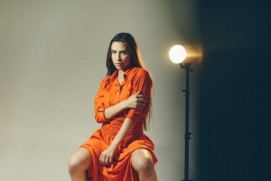 Fashion model seated in an orange shirt dress under a single studio light, the kind of garment-on-body reference shot that AI clothing try-on is judged against